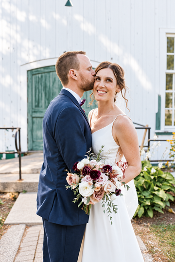 A bride in a sleeveless white wedding gown holding a bouquet of burgundy, cream, and white flowers, standing outdoors next to a groom in a navy blue suit who is kissing her cheek. They are posed in front of a white wooden building with a green door.